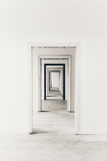 Des portes ouvertes en enfilade, se suivent dans un long couloir - ou peut-être s'agit-il d'un enchainement de pièces vides. Le sol et les murs sont peints en blanc, de même que l'encadrement des portes - sauf une porte bordée de noir.